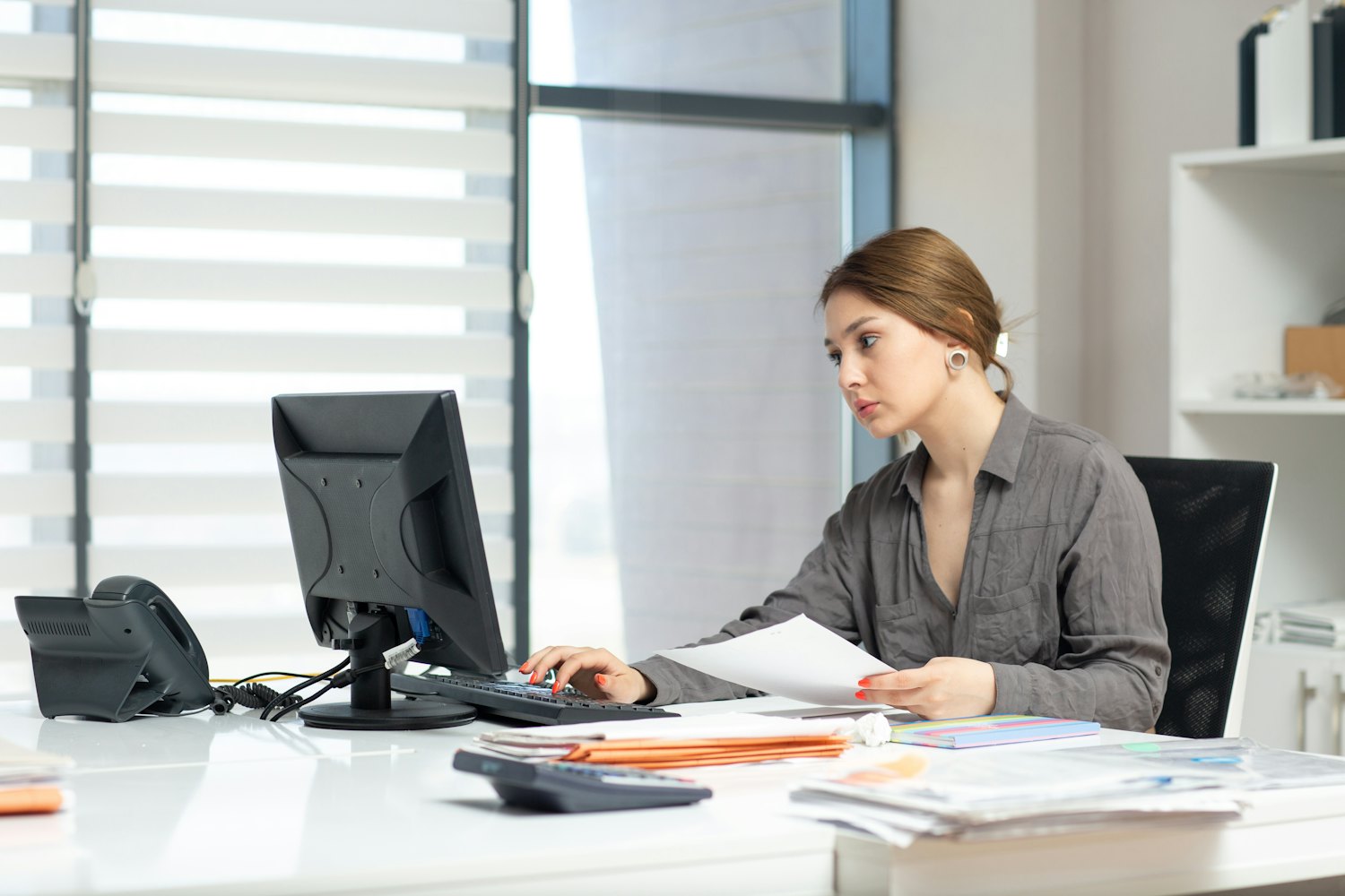 An administrator at a single desk holding a printed document in one hand while keying it into the system on screen with the other.