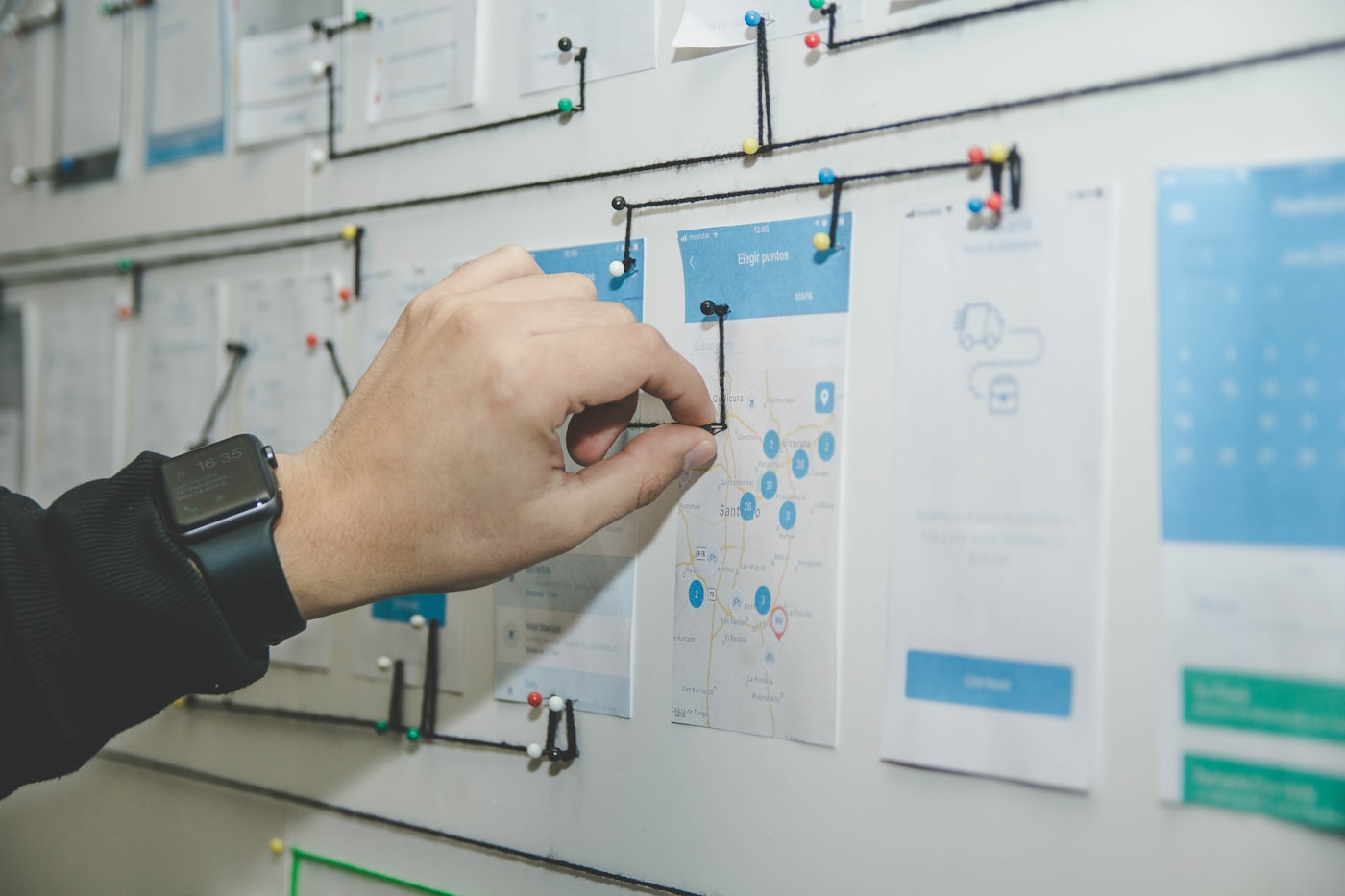 A product flow pinned across a wall as printed app screens joined by string, with a hand moving one of the connections