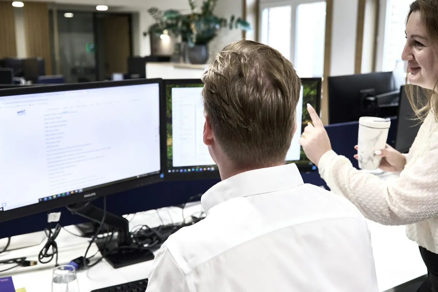 A person at a two-monitor desk working through a long list on screen while a colleague points something out